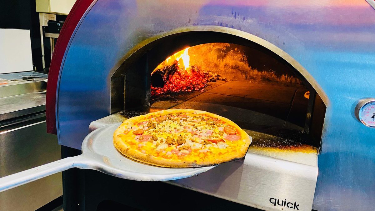 A pizza on a peel in front of Scoozi's lit wood-fired oven.
