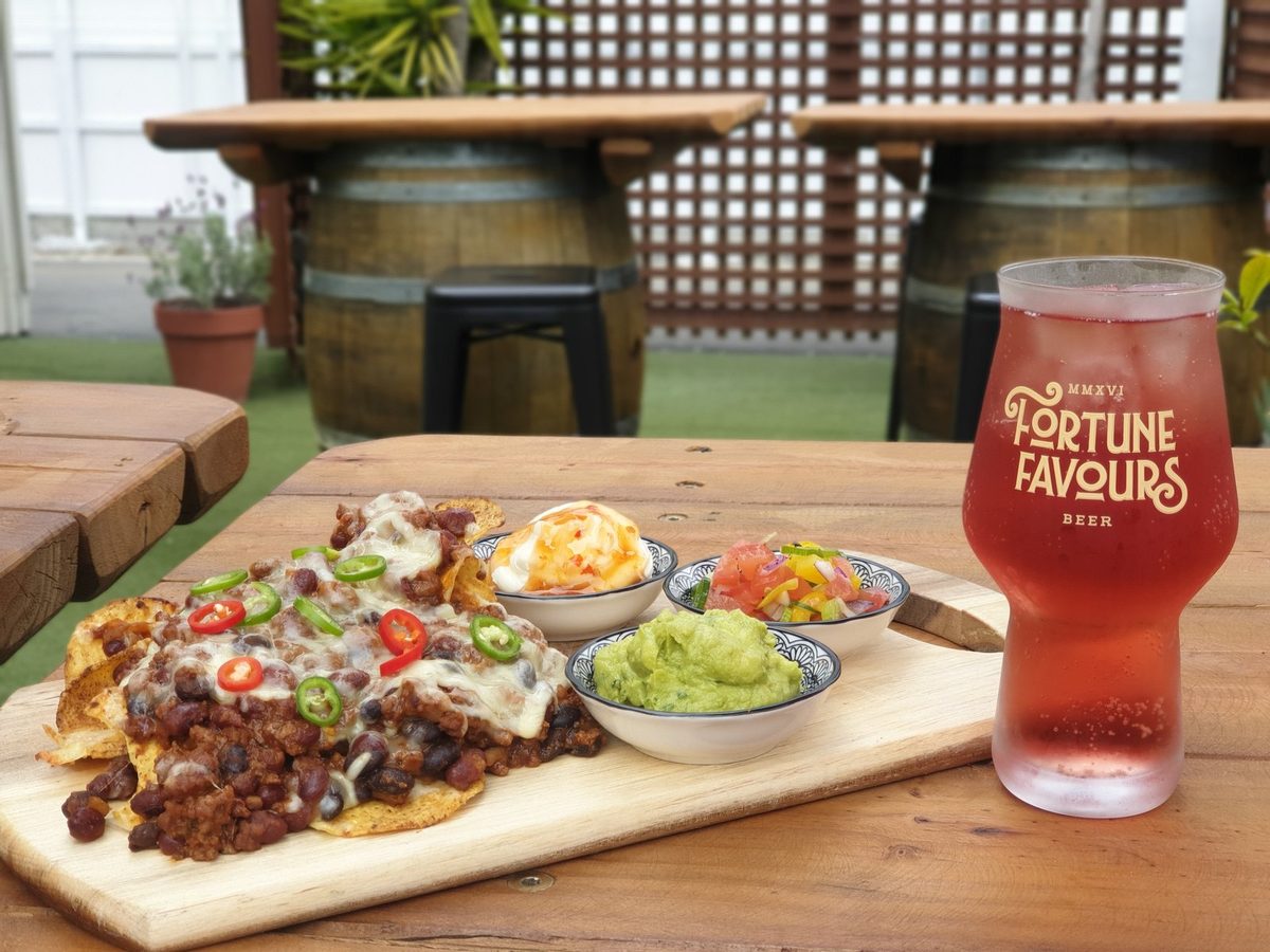 Loaded nachos with guacamole, salsa and sour cream beside a cold red drink in the outdoor seating area.