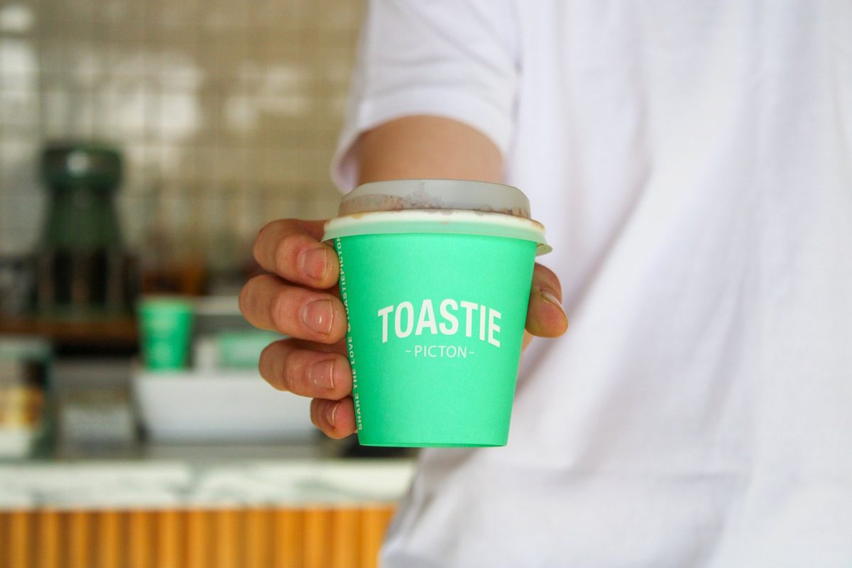 A hot drink in a bright green Toastie Picton branded takeaway cup held over the shop counter.